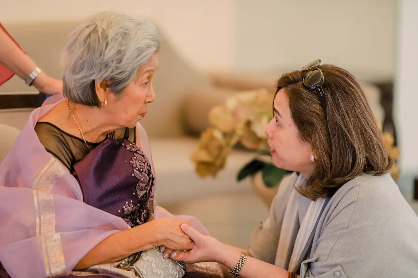 An older woman and her adult daughter holding hands in earnest conversation at home.