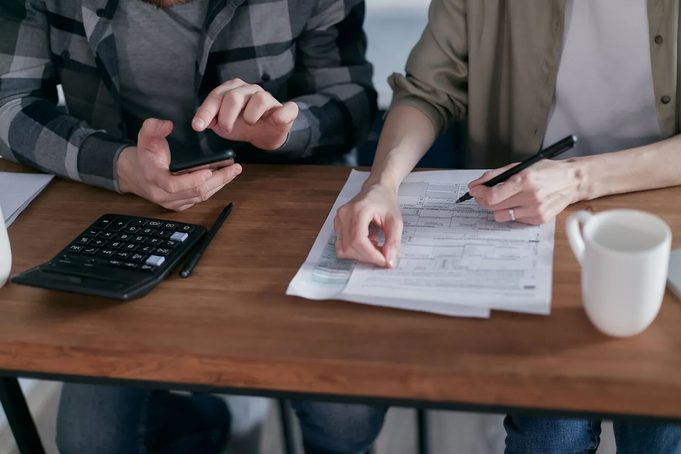 Two people working through figures on paper with a calculator at a wooden desk.