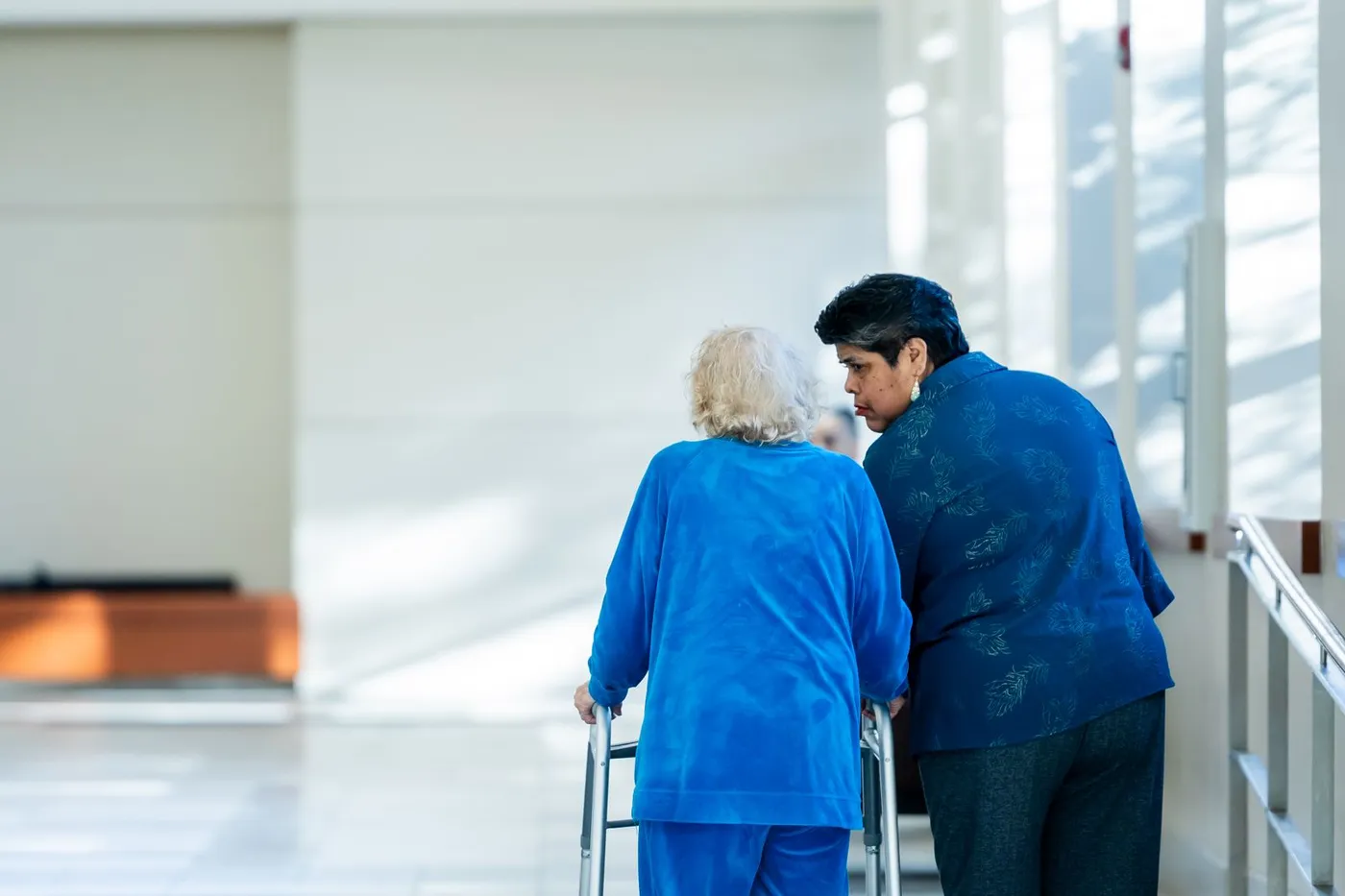 A caregiver helping an elderly woman with a walker in a bright corridor.